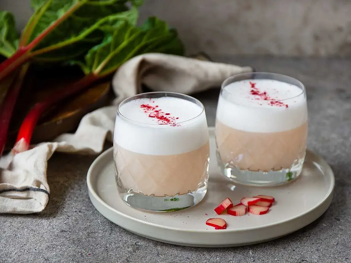 Två glas med en krämig rabarber sour, toppad med skum och strödd med torkad rabarber.