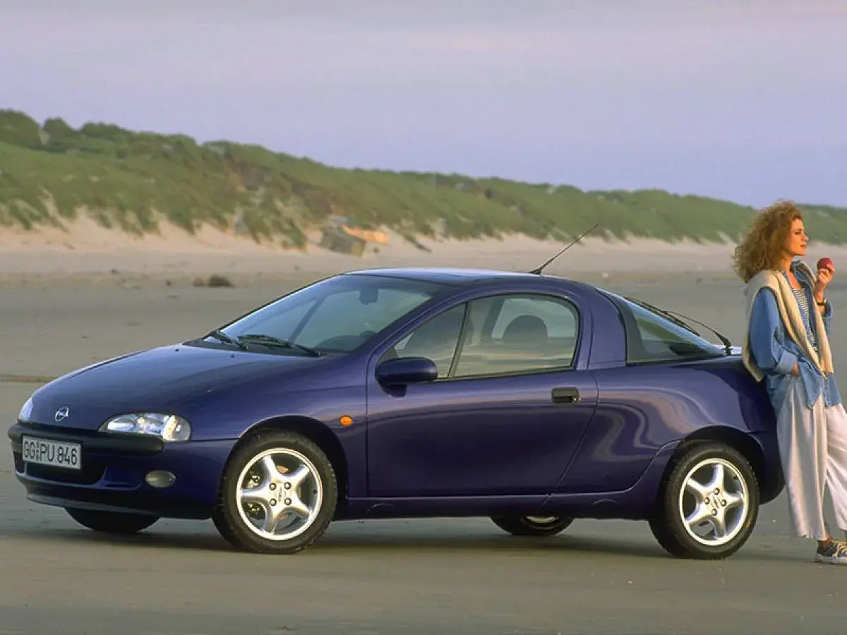 Una donna appoggiata a una Opel Tigra blu, icona delle macchine anni 90, sulla spiaggia al tramonto.