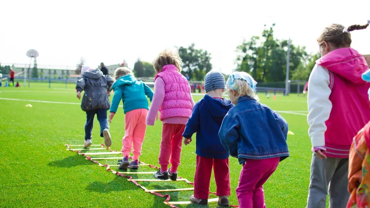 Niños corriendo en una escalera de agilidad en un campo de césped artificial, ideal para aprender inglés en lugares divertidos.