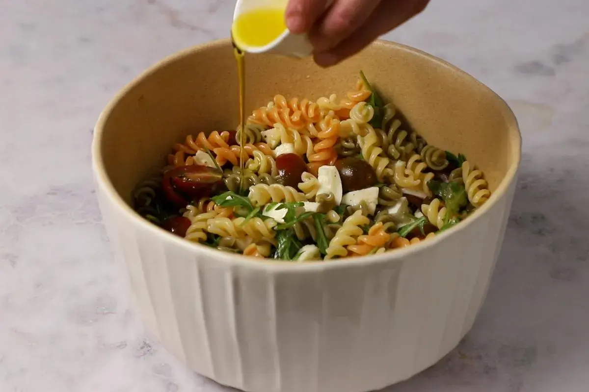 Un bol lleno de pasta de colores, tomates cherry, queso y rúcula, con aceite de oliva vertiéndose sobre ella.