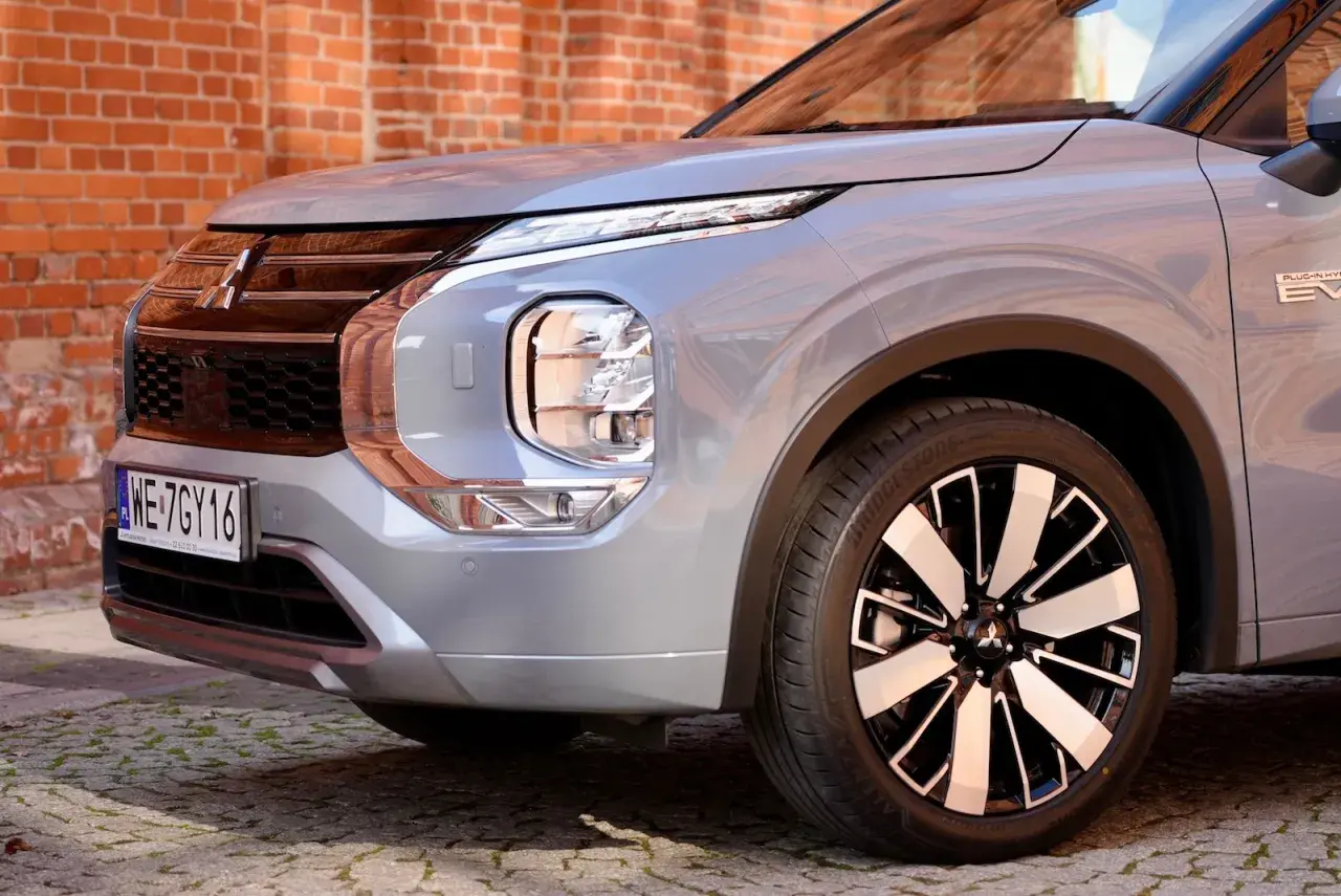 Przód srebrnego Mitsubishi Outlander 2025 z charakterystycznym grillem i reflektorami LED, zaparkowany na bruku przed ceglaną ścianą.