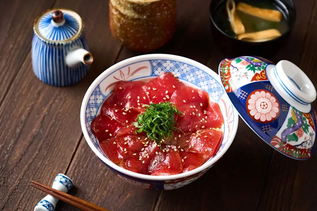A bowl of delicious Japanese sweet rice topped with thinly sliced tuna, sesame seeds, and green garnish. A decorative lid rests beside it.