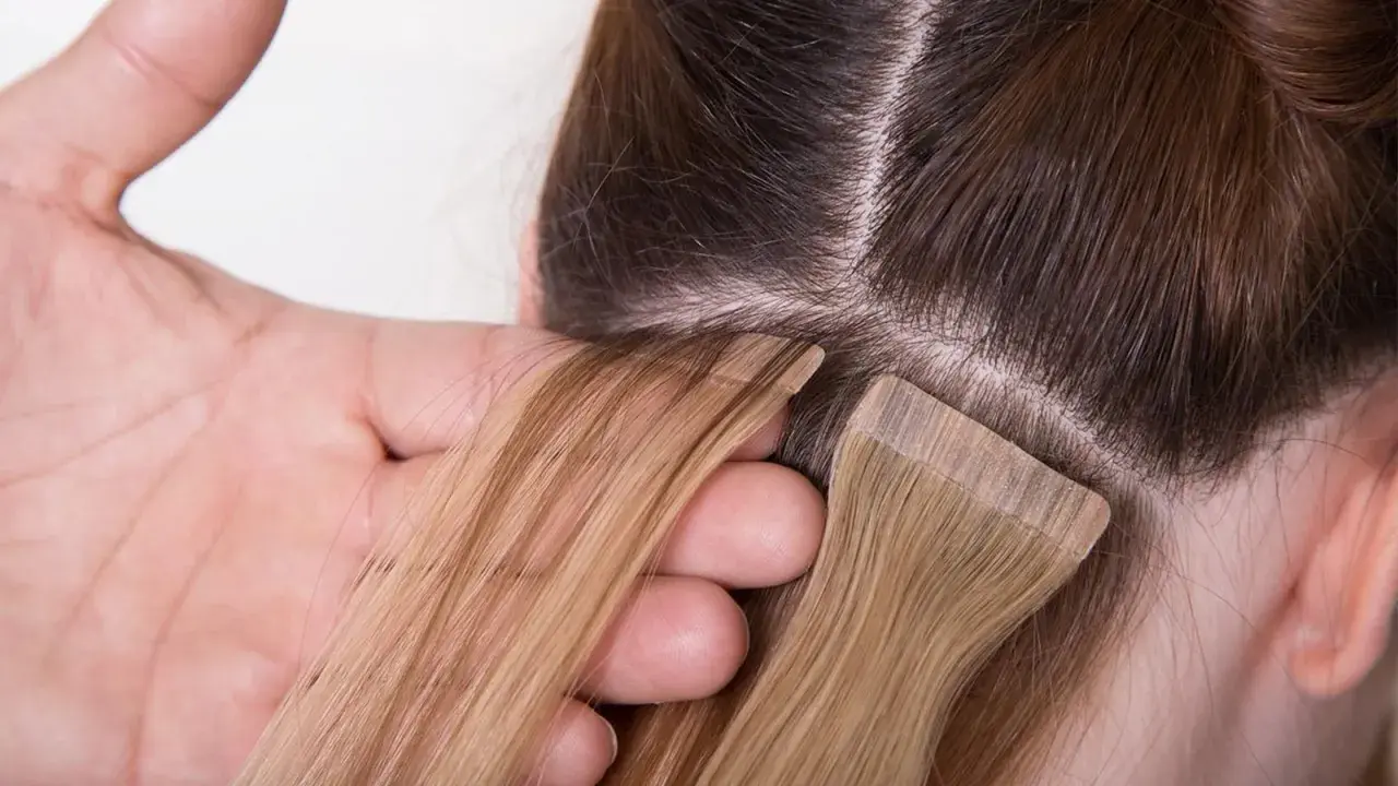 Manos aplicando extensiones adhesivas rubias a cabello castaño. El proceso de colocación de extensiones adhesivas es delicado.