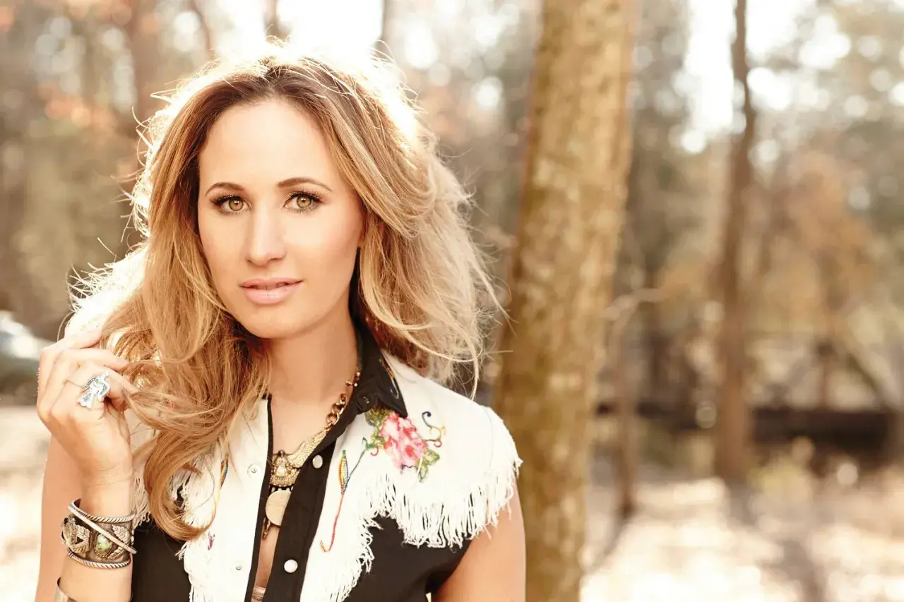 Jessi Alexander, a blonde woman with striking eyes, poses outdoors in a fringed vest adorned with floral embroidery.