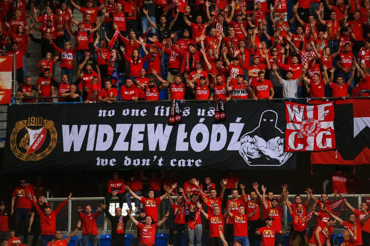 Tłum kibiców Widzewa w czerwonych koszulkach skanduje przyspiewki na Widzew, machając rękami. Na banerze widnieje napis "WIDZEW ŁÓDŹ".