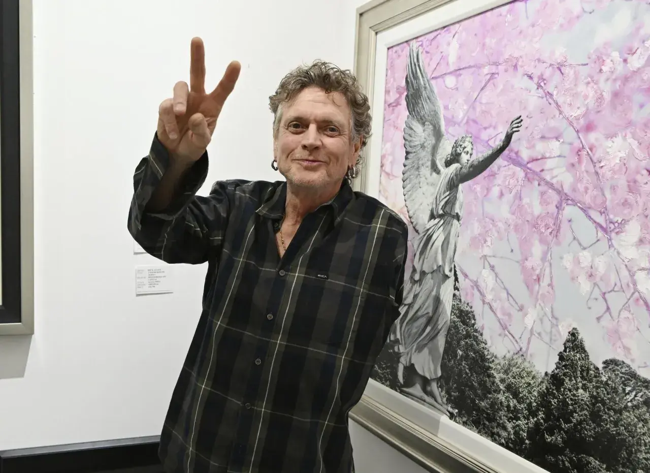 Rick Allen, perkusista Def Leppard, pozuje z uniesionym znakiem pokoju obok obrazu z aniołem i kwitnącymi drzewami.