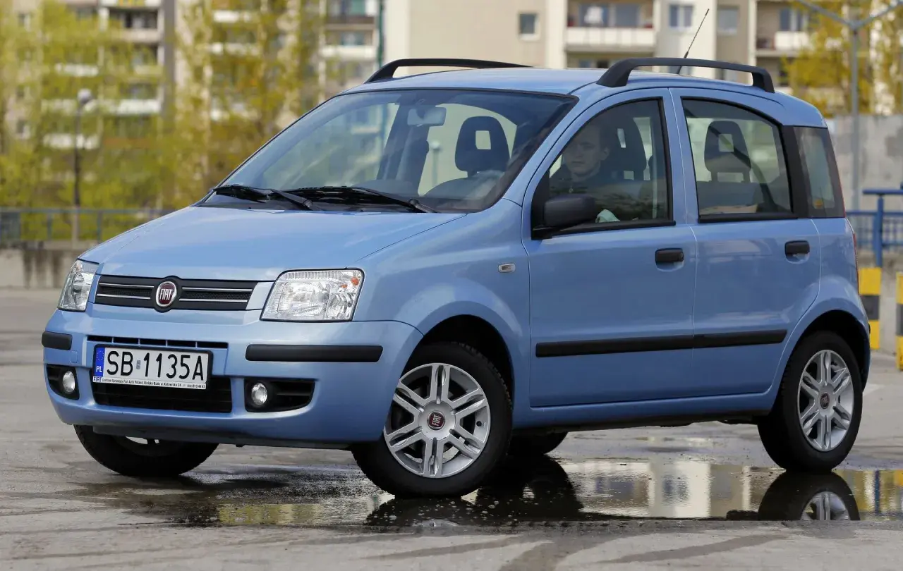 Niebieski Fiat Panda z kierowcą, stojący na mokrej nawierzchni. Kierowca zastanawia się nad aktualną ceną gazu.
