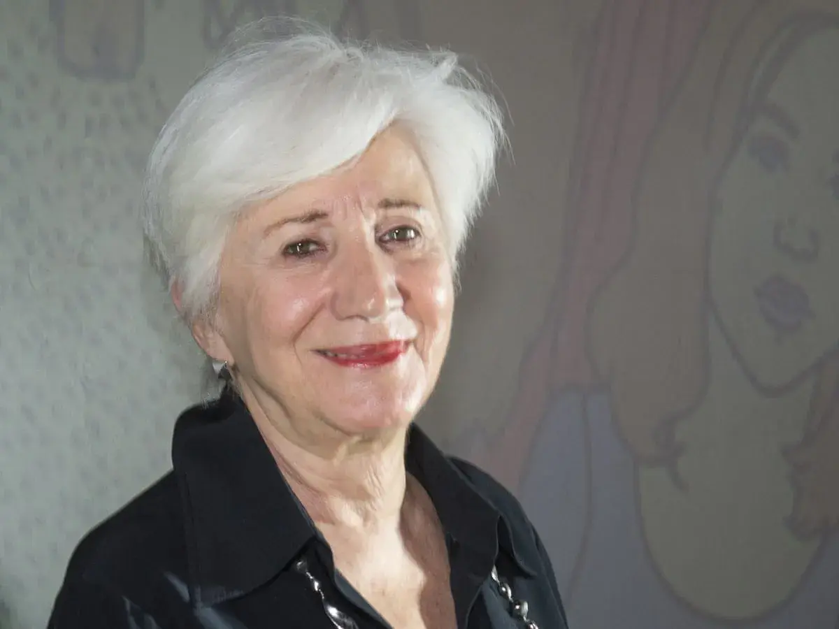 Olympia Dukakis ler vänligt mot kameran, med sitt karakteristiska vita hår och en svart skjorta. Bakom henne anas en tecknad figur.