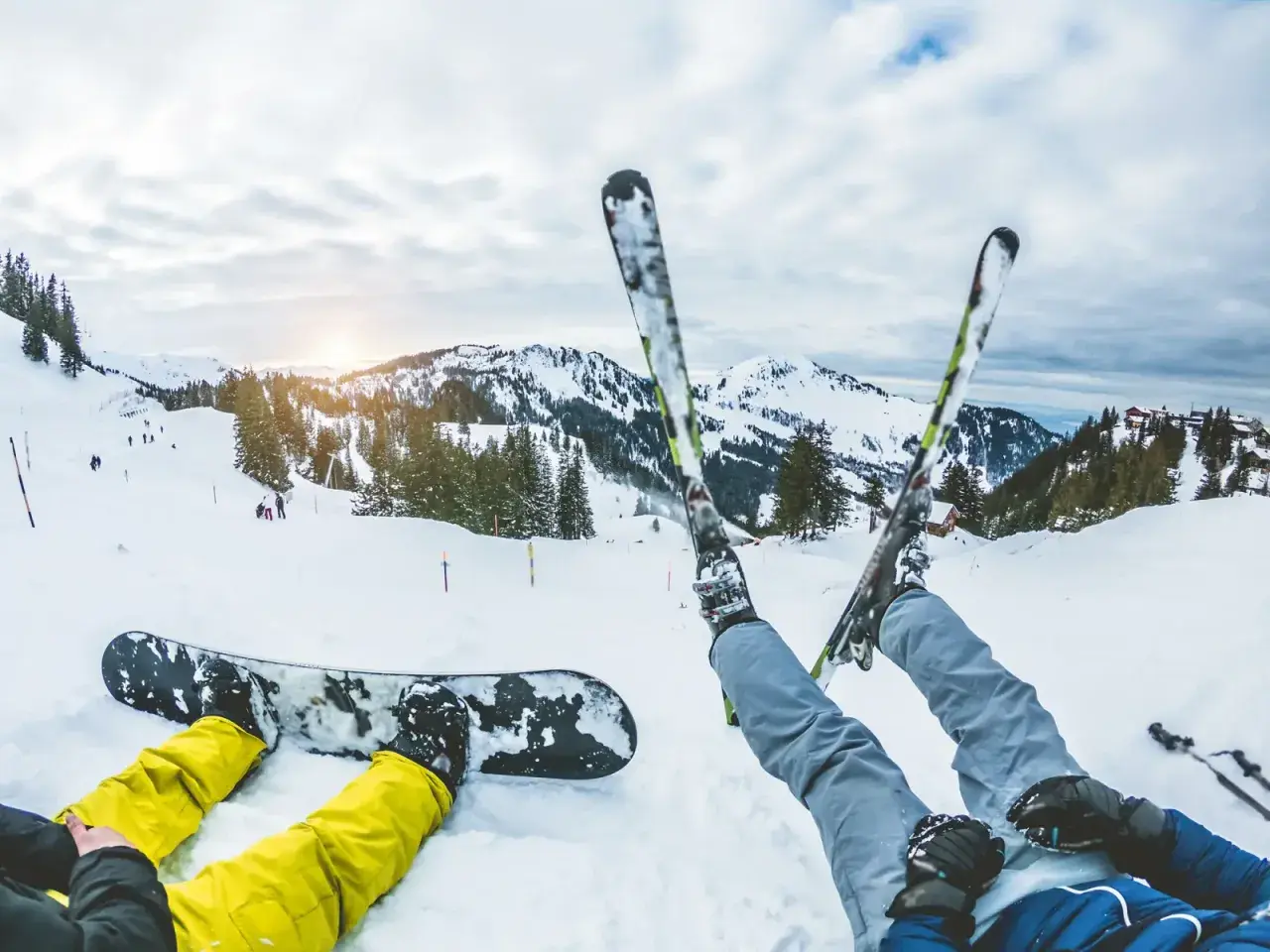 Dwie osoby odpoczywają na śniegu. Jedna leży na desce snowboardowej, druga ma założone narty. W tle góry i stoki narciarskie.