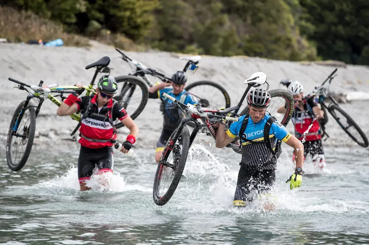 Teilnehmer eines **adventure racing** durchqueren mit Fahrrädern auf den Schultern einen Fluss.