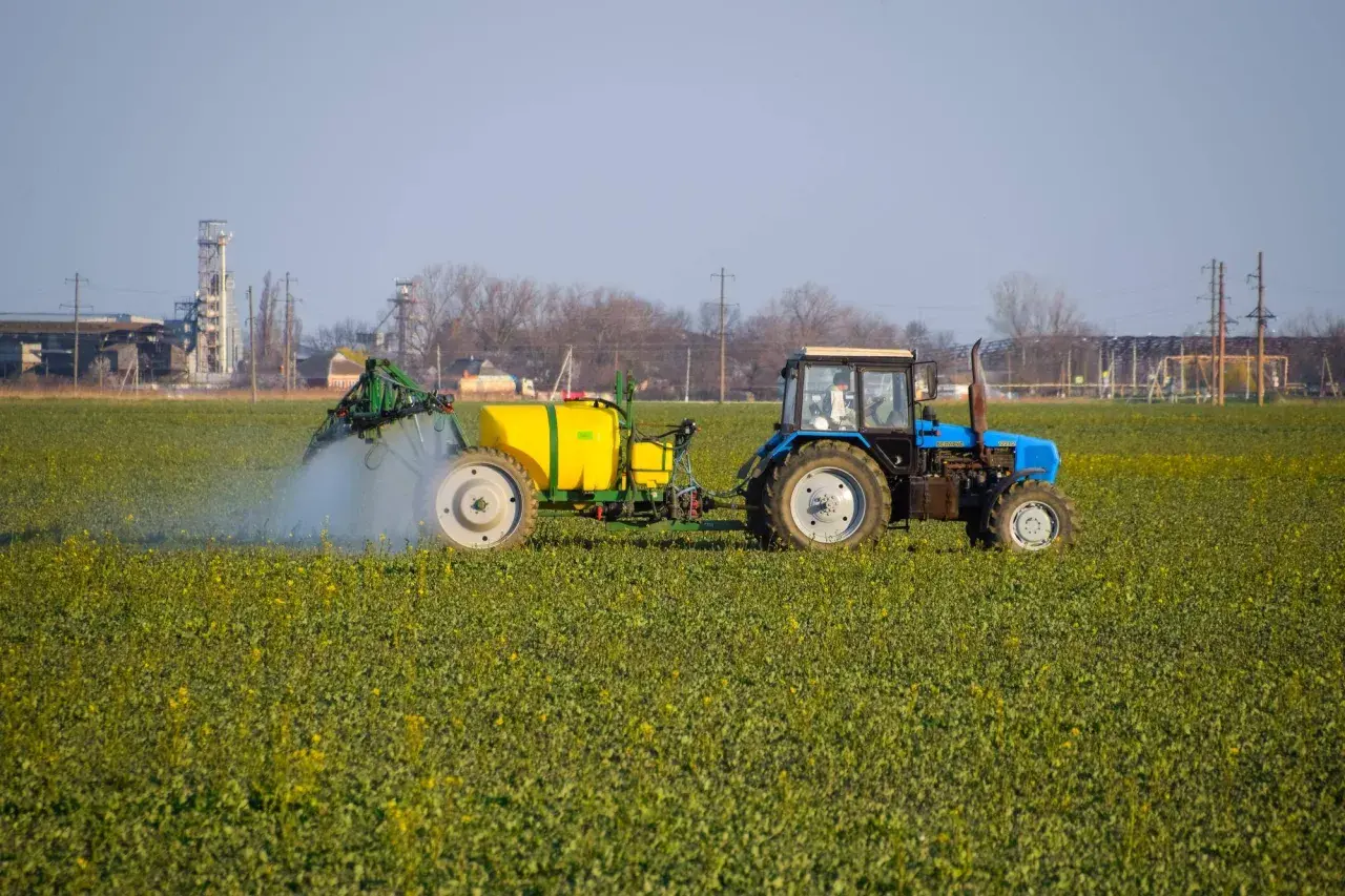 Niebieski traktor z żółtym zbiornikiem wykonuje oprysk na polu zielonych roślin. W tle widać przemysłowe budynki.