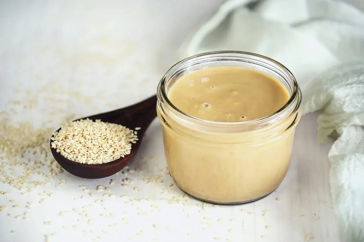 Un vasetto di tahina, crema di sesamo, accanto a un cucchiaio di semi. Scopri cos'è la tahina e come usarla.
