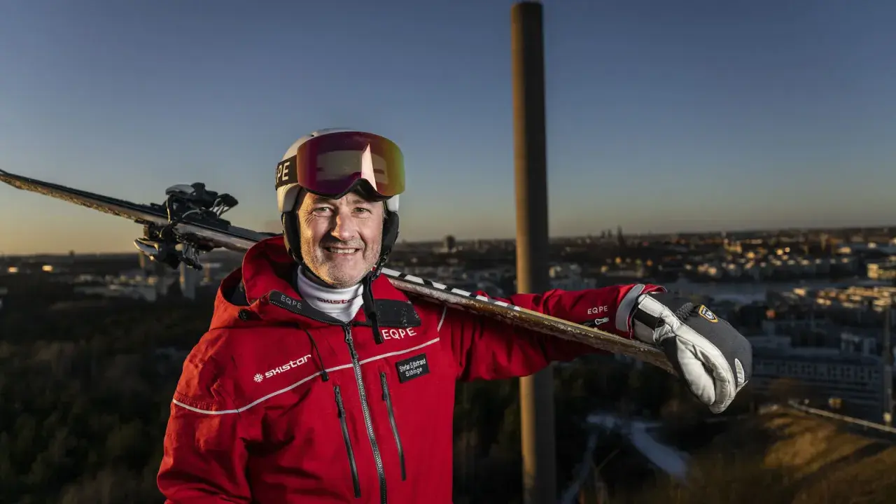Ski-entusiast med skidor på axeln, klädd i röd jacka med SkiStar-logga. Han ser ut att vara en del av SkiStar ledning, redo för äventyr.