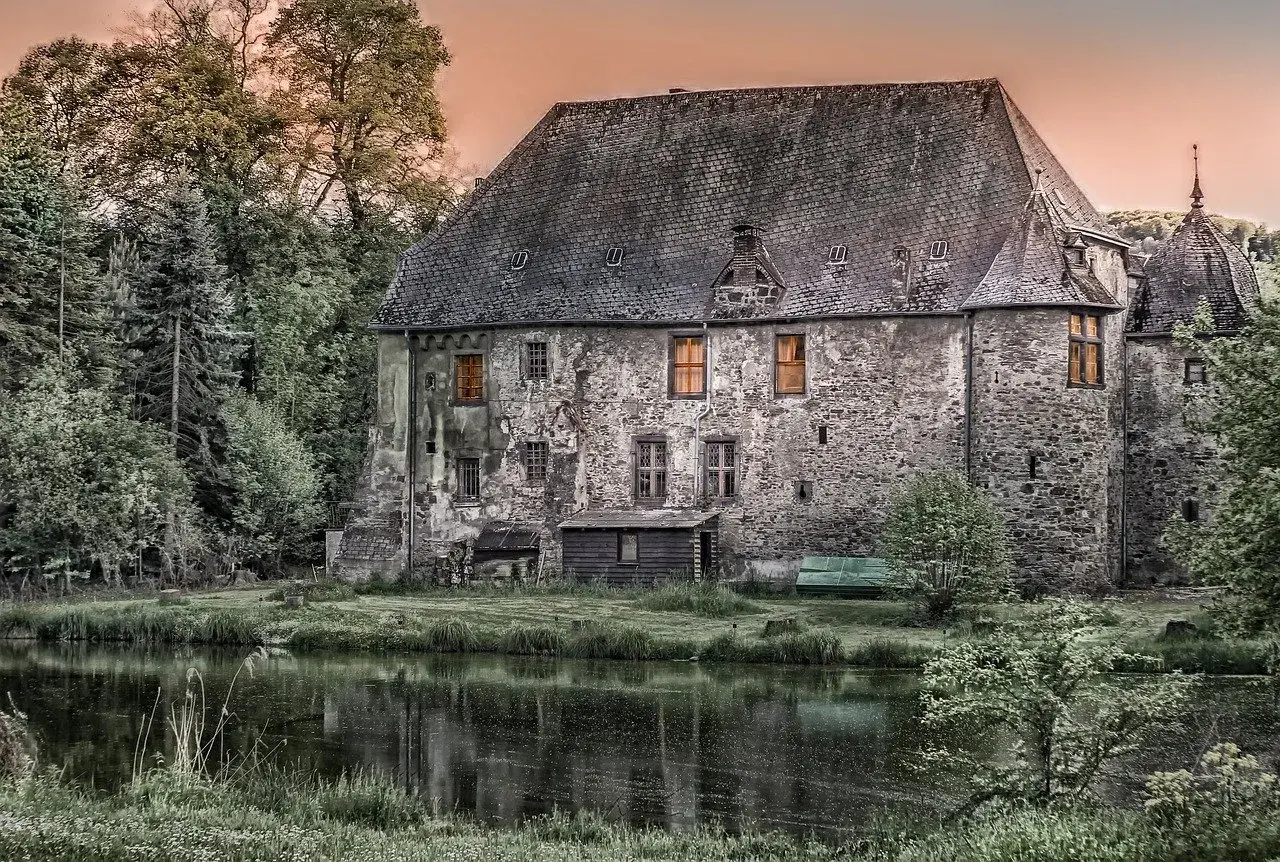 Kamienny polski dwór szlachecki z oknami oświetlonymi ciepłym blaskiem, otoczony zielenią i stawem.