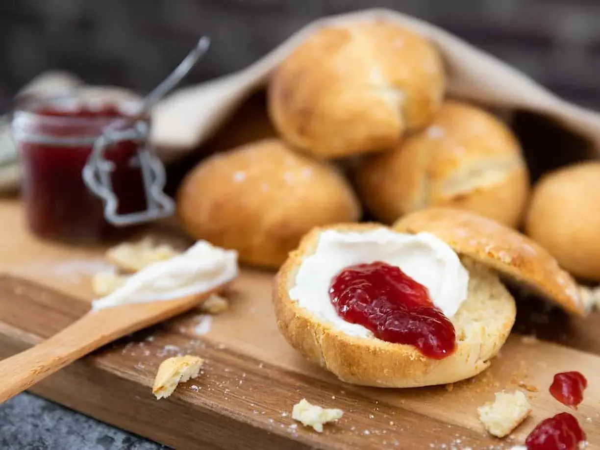 En sked med grädde och sylt på en halverad scones, redo att avnjutas. En burk sylt och fler av dessa bästa scones i bakgrunden.