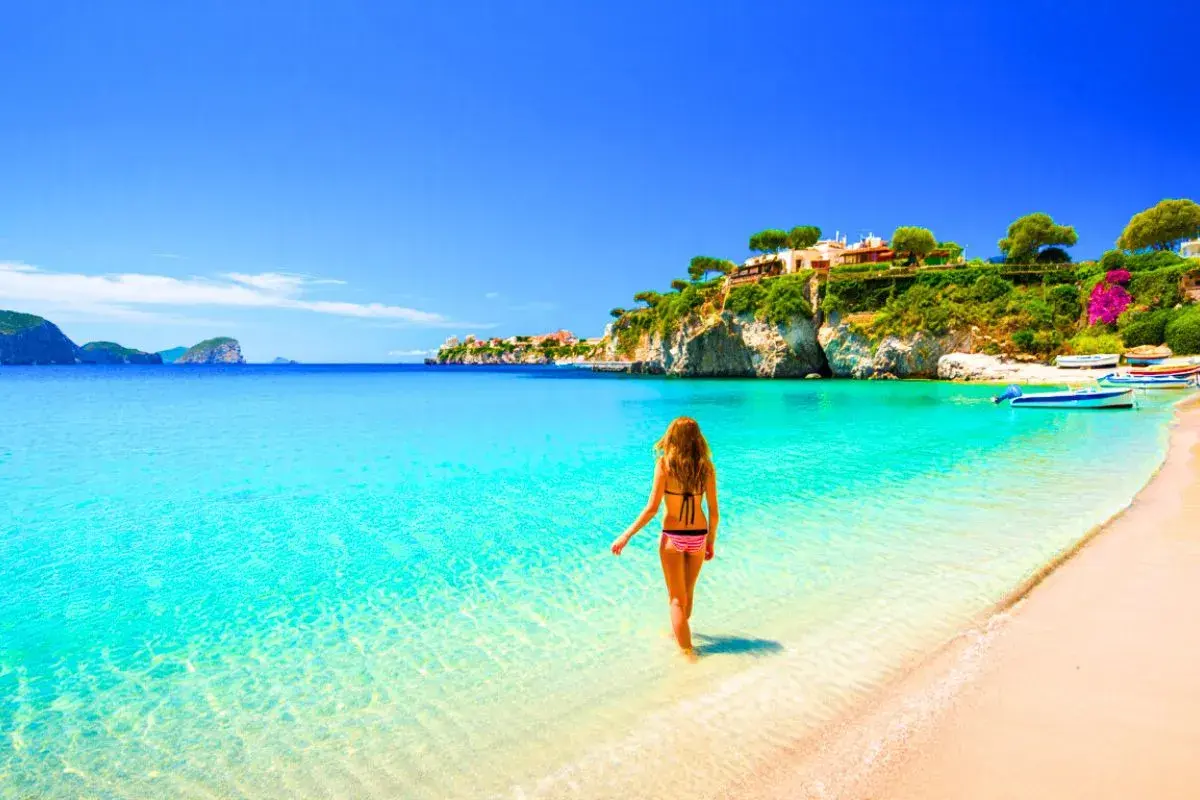 Femme en bikini entrant dans l'eau turquoise d'une plage idyllique, avec des maisons perchées sur une falaise, typique de la côte méditerranée en Italie.