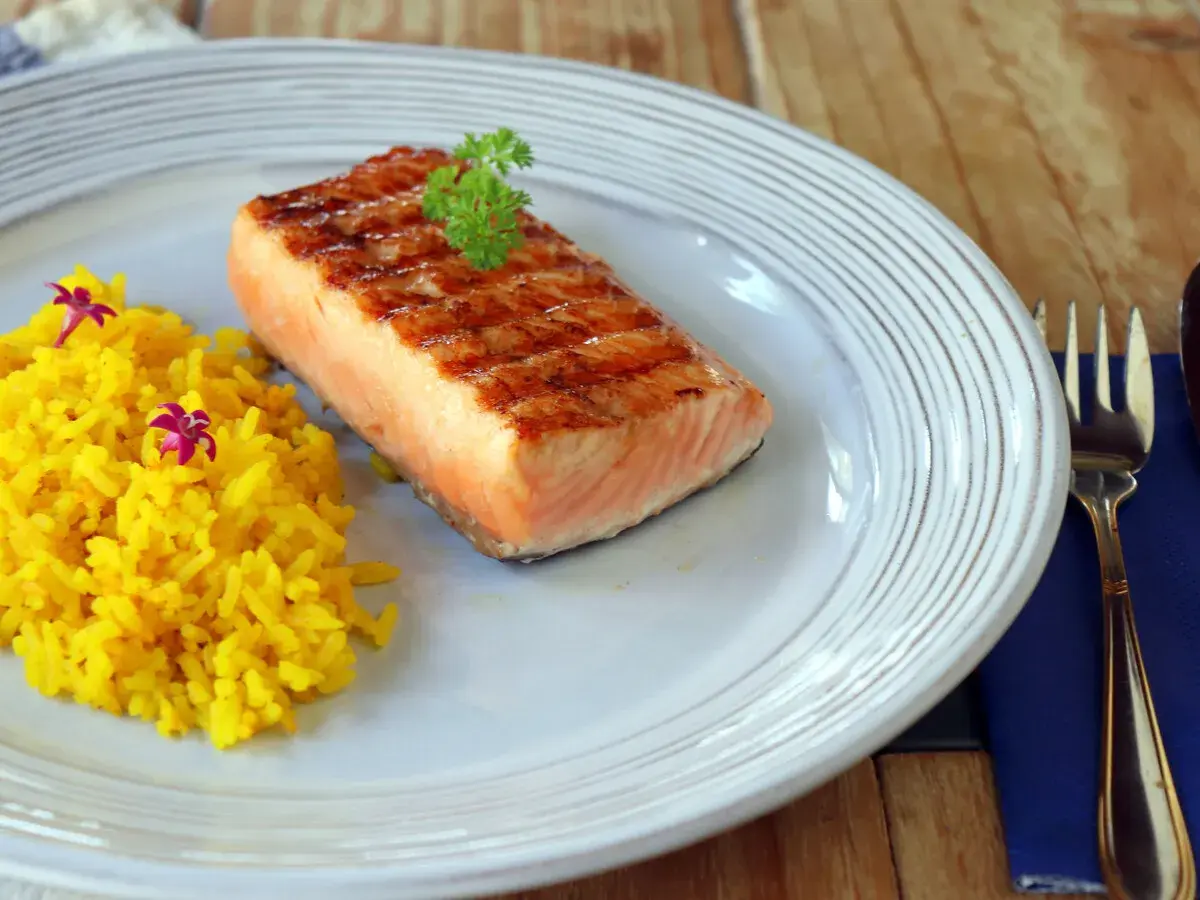 Un delicioso salmón a la plancha con marcas de parrilla, servido con arroz amarillo y decorado con perejil y flores moradas.