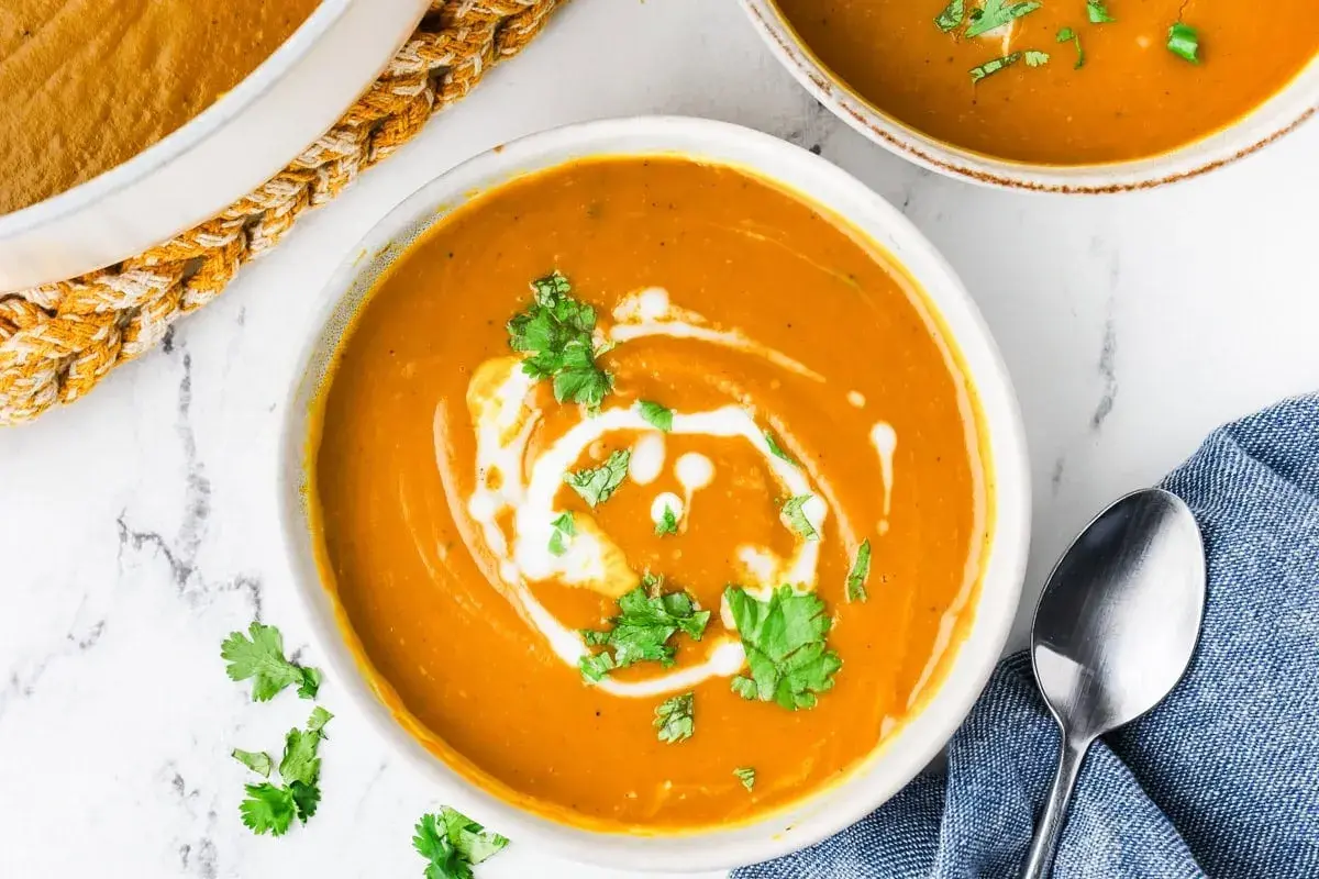 A creamy, vibrant bowl of kabocha squash soup, swirled with coconut cream and garnished with fresh cilantro. A spoon rests on a blue napkin beside it.