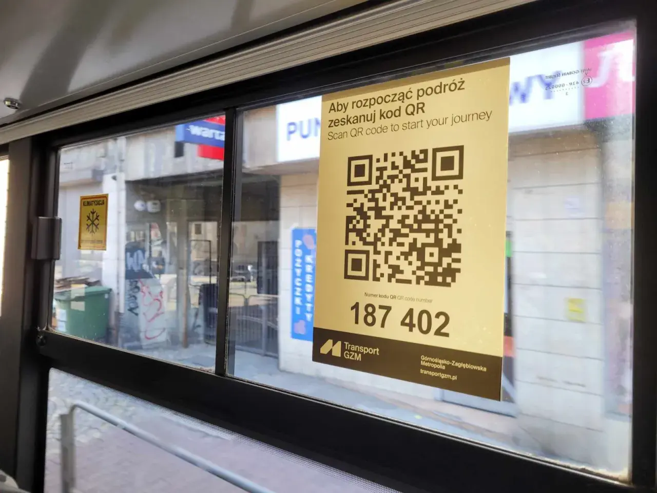 W autobusie widoczny jest żółty plakat z kodem QR i napisem "Aby rozpocząć podróż zeskanuj kod QR". Pod kodem znajduje się numer 187 402.