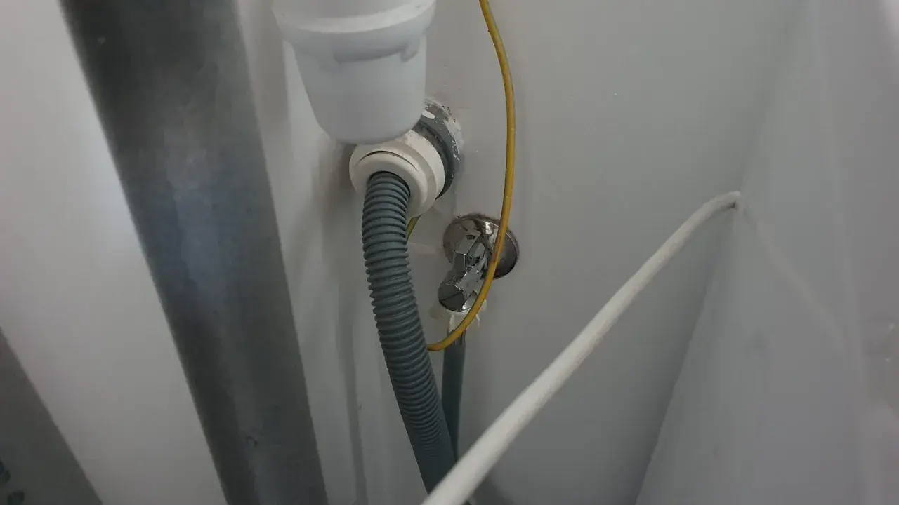 Szara rura odpływowa pralki podłączona do syfonu pod zlewem. Widoczny żółty przewód i biały kabel.