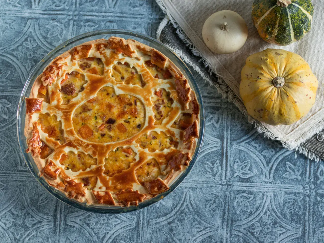 Torta salata zucca salsiccia e stracchino, con decorazione a intreccio e ripieno dorato. Accanto, zucche decorative.