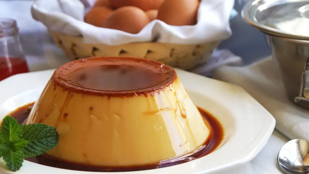 Delicioso flan de huevo casero con caramelo líquido, decorado con menta fresca. En el fondo, una cesta con huevos y un frasco de mermelada.