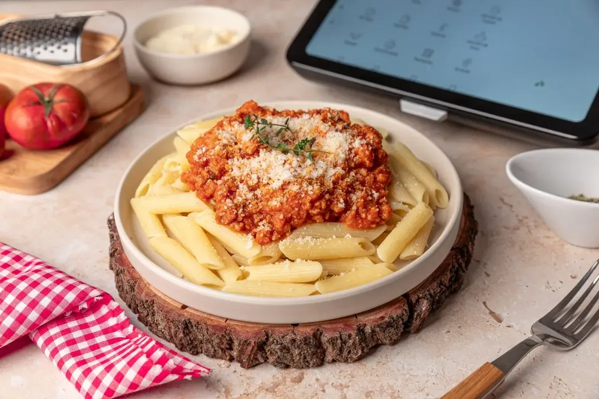 Plato de macarrones a la boloñesa, con queso rallado y hierbas frescas. Ingredientes frescos y una tablet con recetas en el fondo.