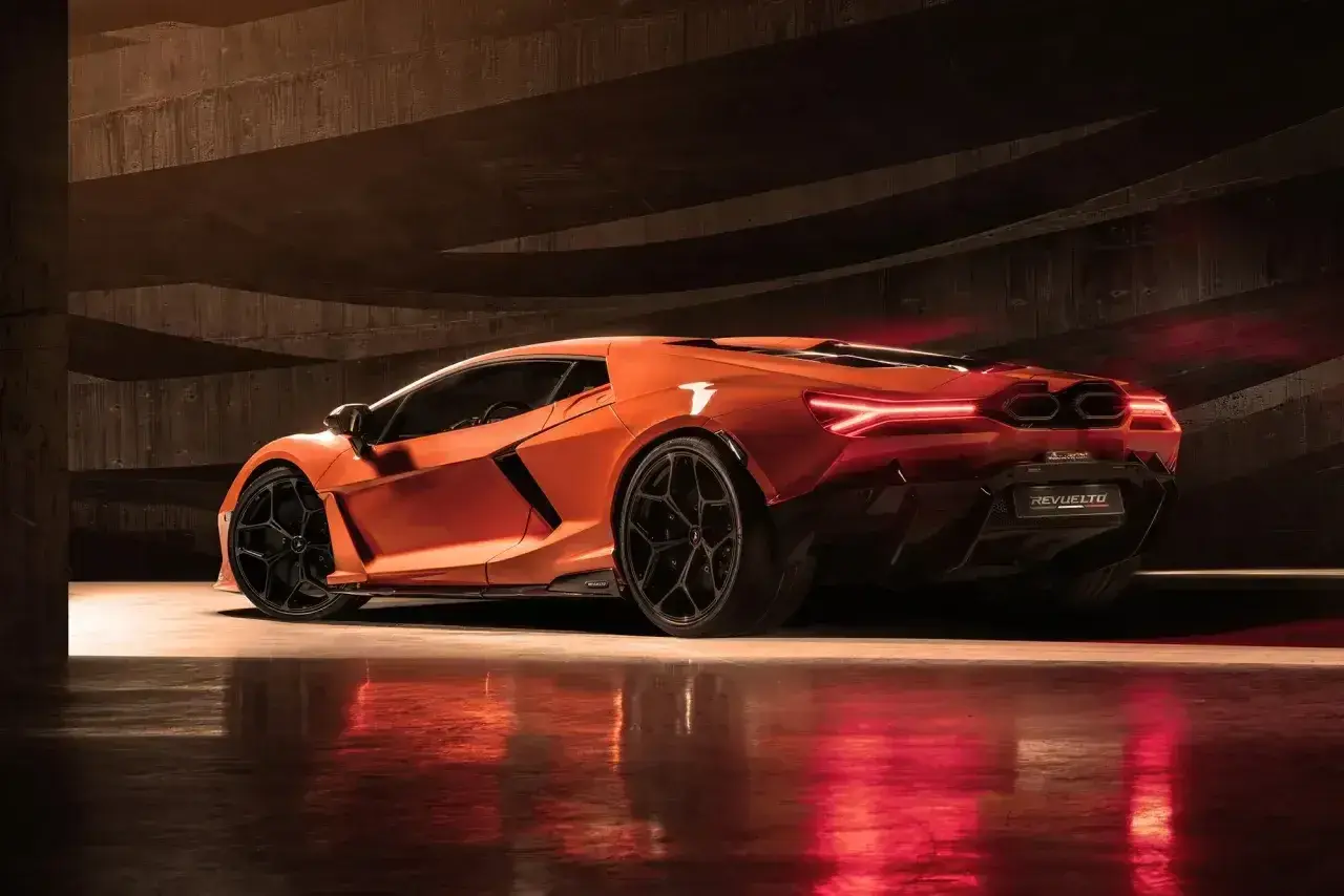 Pomarańczowy Lamborghini Revuelto z czarnymi felgami, stojący w ciemnym, betonowym garażu, z czerwonymi światłami odbijającymi się od podłogi.