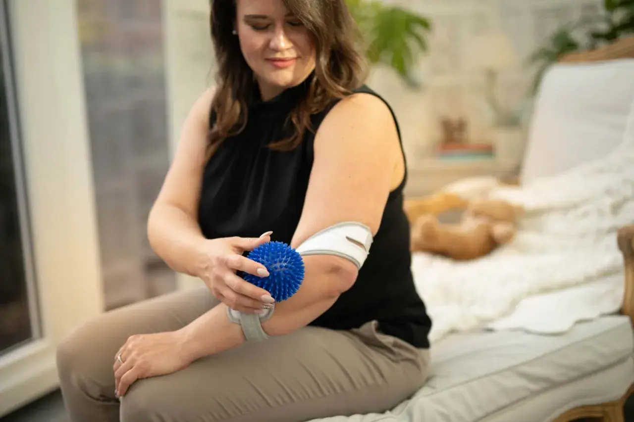 Frau massiert ihren Unterarm mit einem blauen Igelball und trägt eine Bandage.