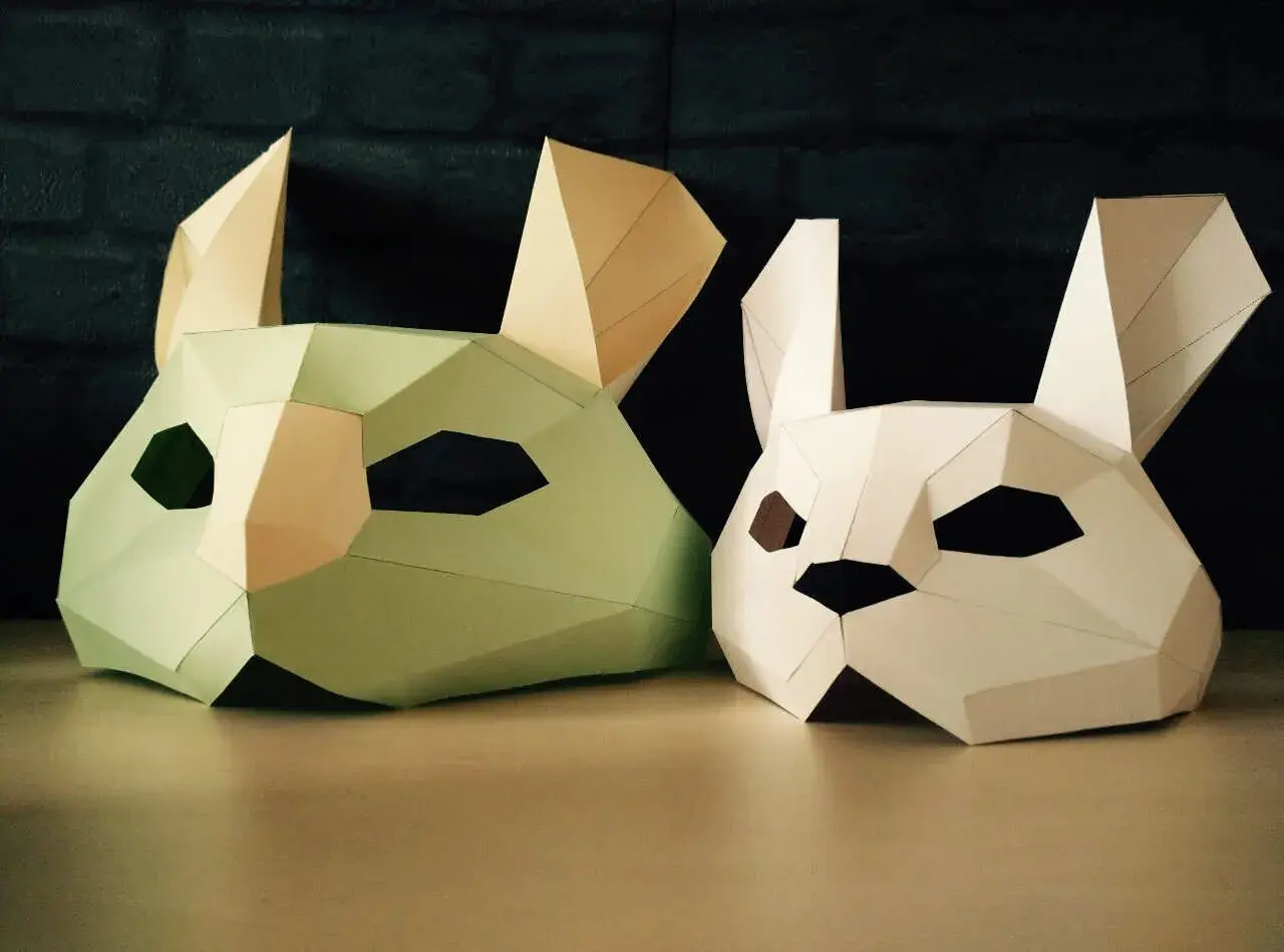 Två påskpyssel i papper: en ljusgrön och en vit kaninmask med stora öron och geometriska former.