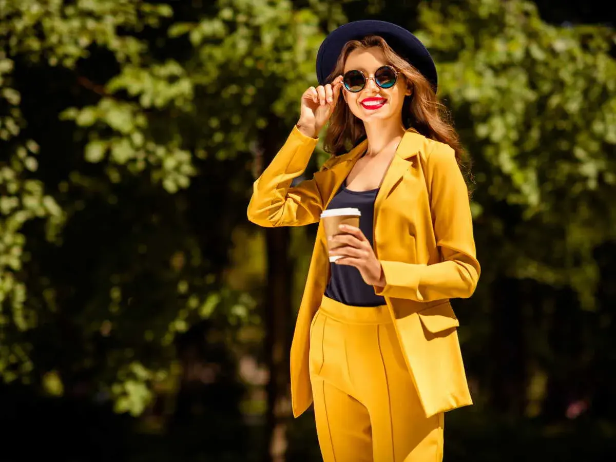 Une femme en costume jaune vif, chapeau et lunettes de soleil, tenant un café. Parfait pour savoir comment s'habiller quand il fait 14 degrés.