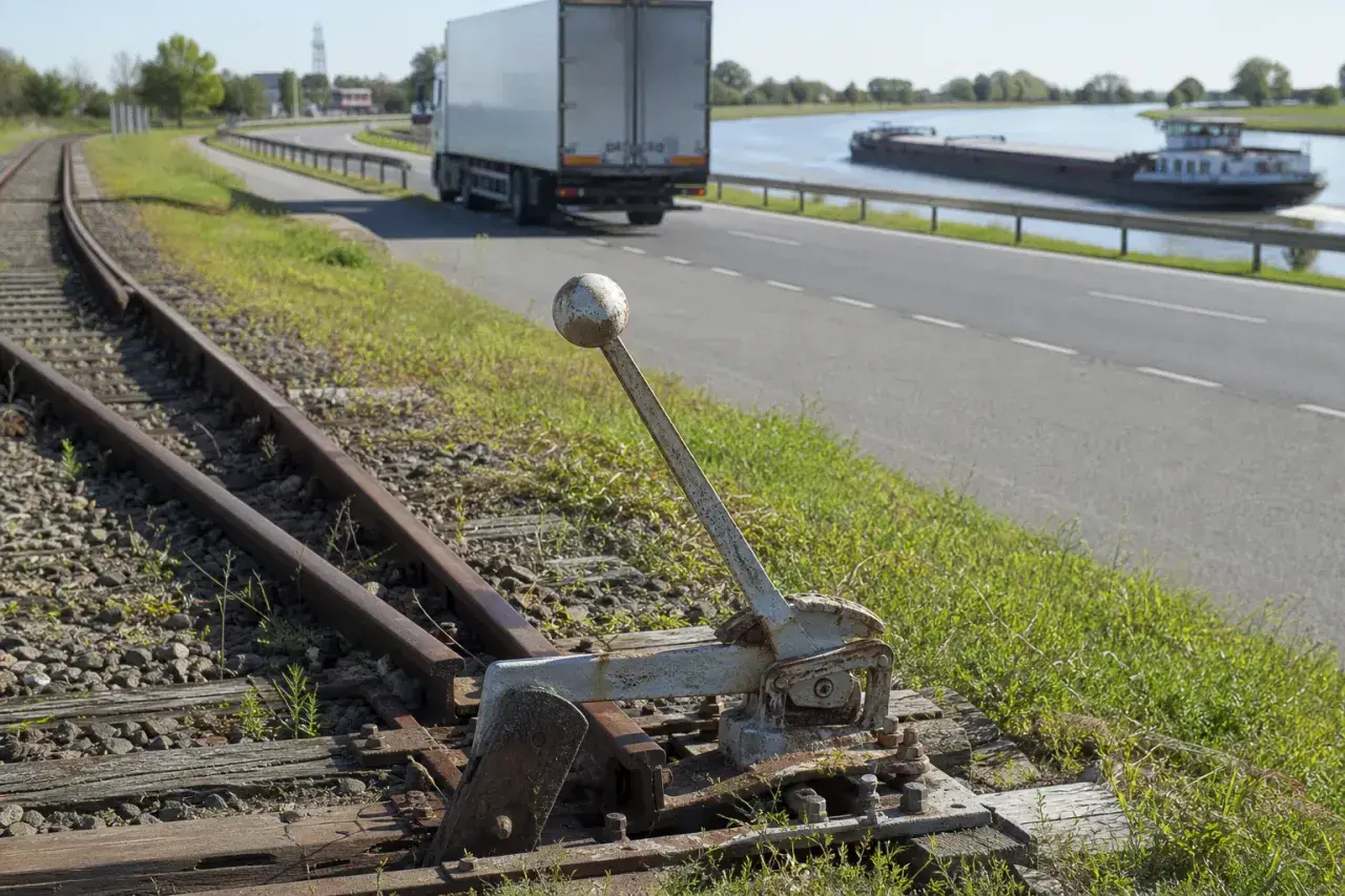 Alte Weiche neben Gleisen. Ein LKW und ein Frachtschiff sind auf der Straße und dem Wasser zu sehen, verschiedene Verkehrsmittel.