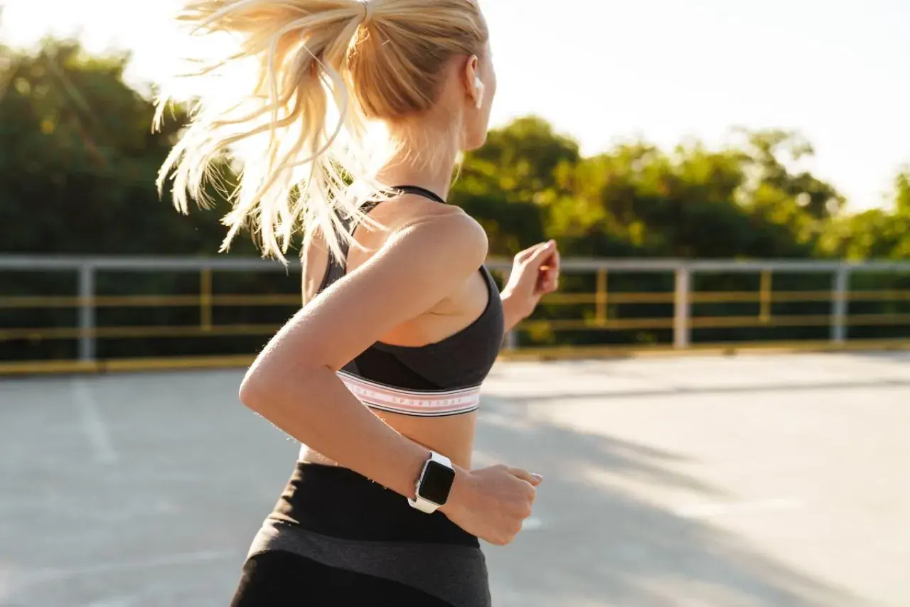 Kobieta w sportowym stroju wykonuje ćwiczenia cardio, biegając na świeżym powietrzu. Słońce rozświetla jej blond włosy.