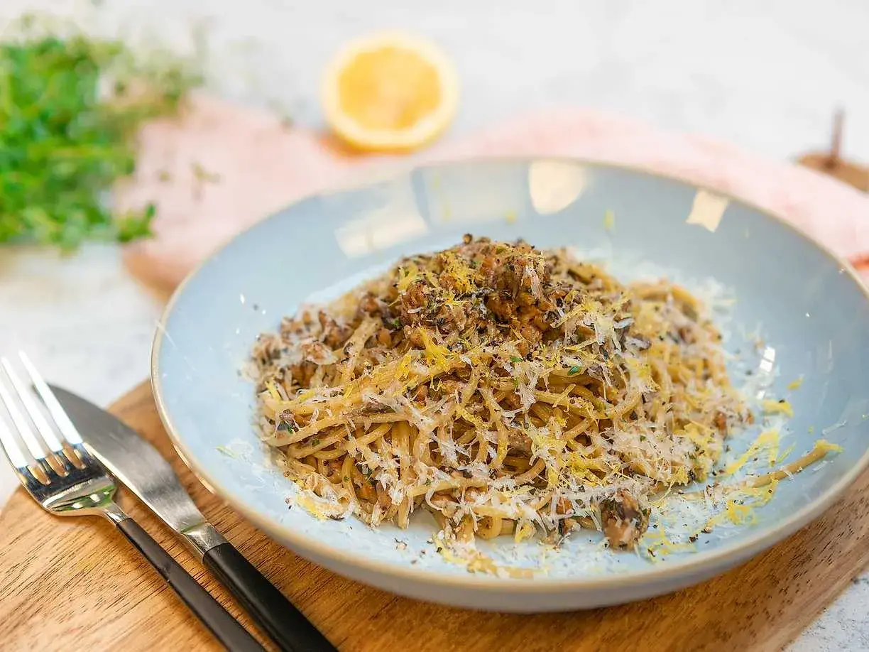 En tallrik med saffranspasta vegetarisk, toppad med riven ost och citronzest. Serveras med bestick på en träskärbräda.