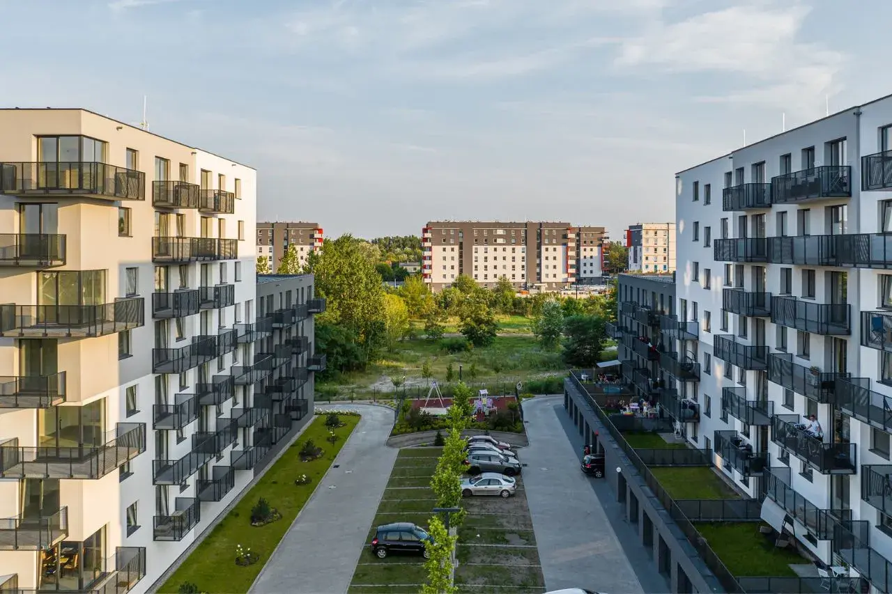 Nowoczesne osiedle mieszkaniowe, z białymi blokami i balkonami, otoczone zielenią. W tle widać kolejne budynki, tworzące krajobraz jak z **okam capital**.