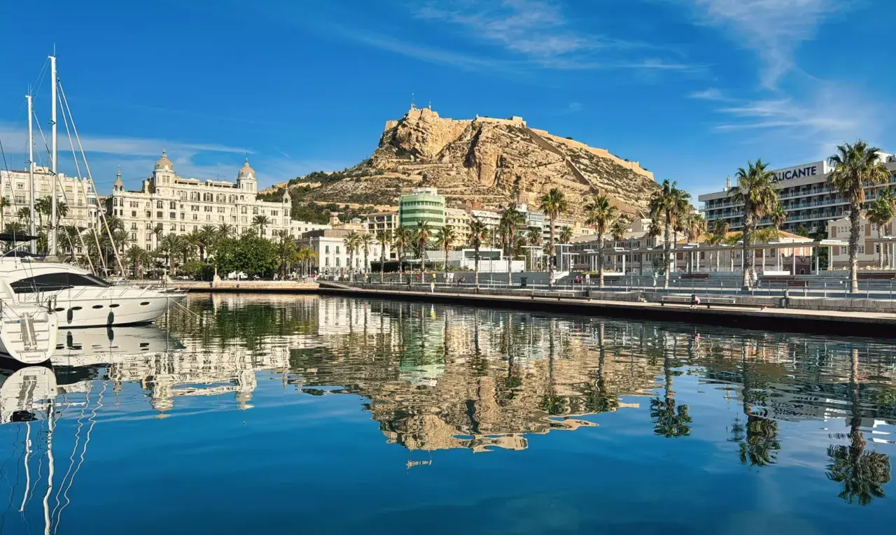 Alicante, luty. Słoneczny dzień nad zatoką z odbiciem zamku Santa Barbara i palm w krystalicznie czystej wodzie.