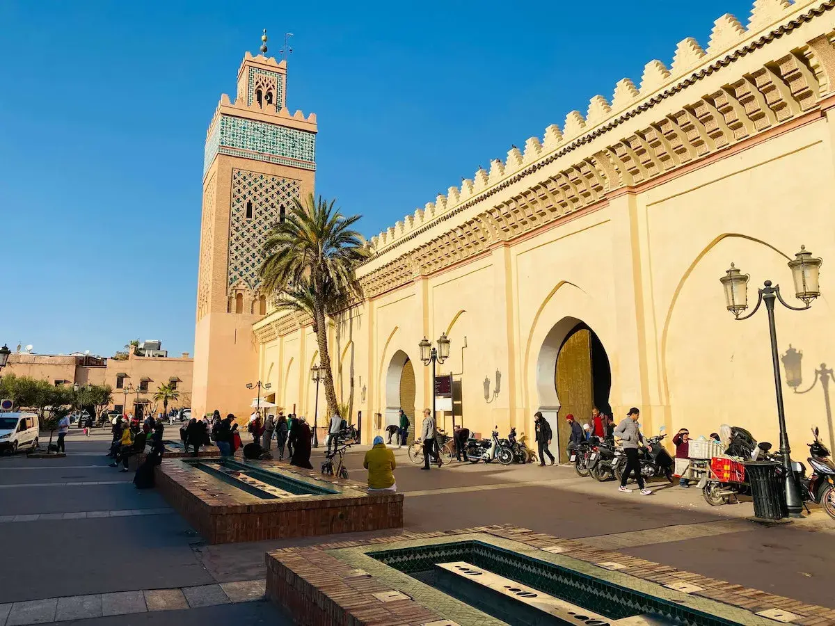 La moschea Koutoubia domina la piazza, con la sua torre decorata e le arcate del souk di Marrakech. Gente e motorini animano la scena.