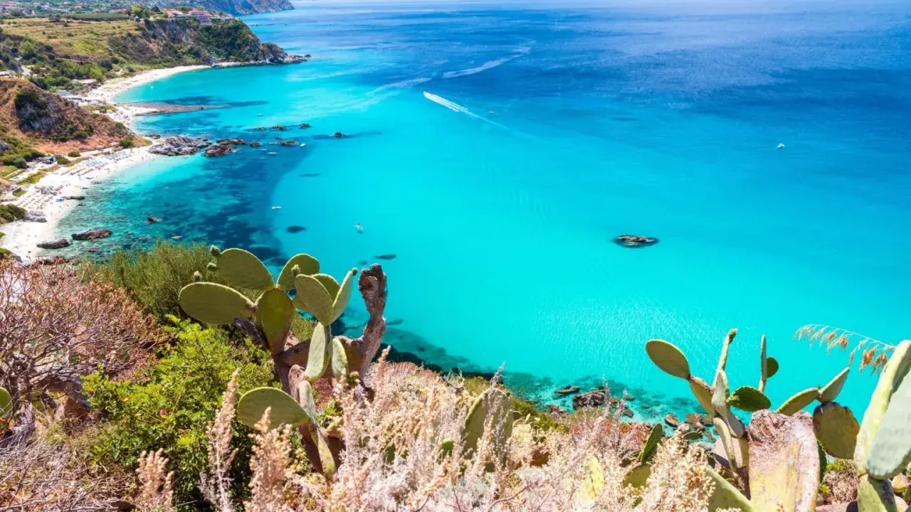 A stunning view of Reggio Calabria beaches, with turquoise waters, white sand, and lush green cliffs dotted with cacti.