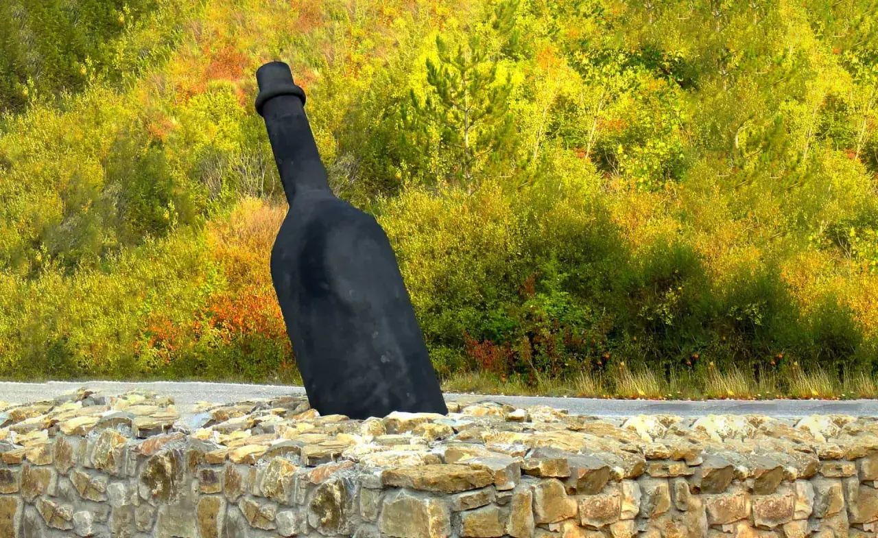 Giant black wine bottle sculpture emerging from a stone wall, with a colorful autumn forest in the background.