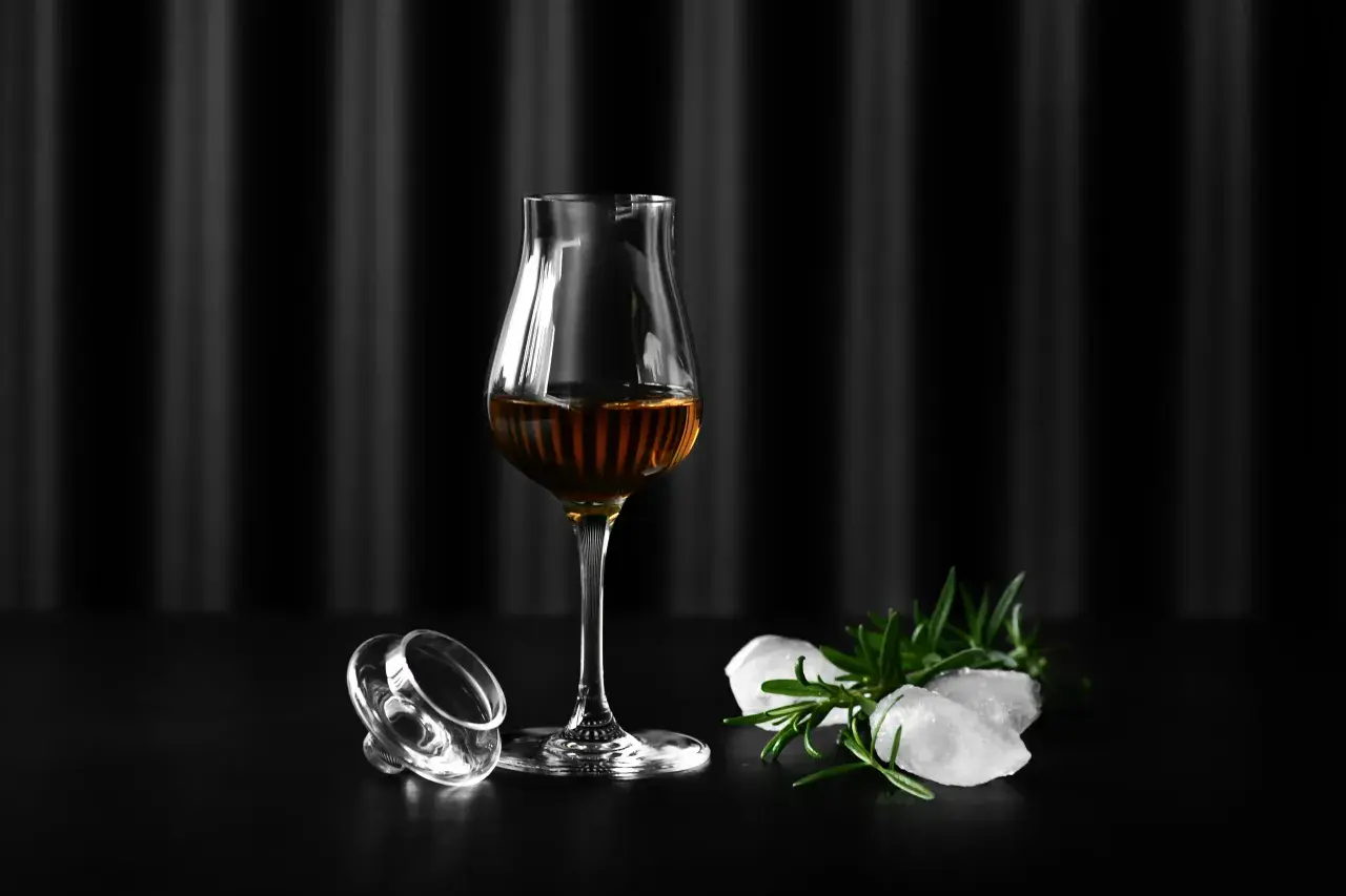 Szklanka Glencairn z bursztynowym trunkiem, obok leży jej zakrętka, lód i rozmaryn. Jak dobrać odpowiednie szkło Glencairn vs tumbler vs copita.