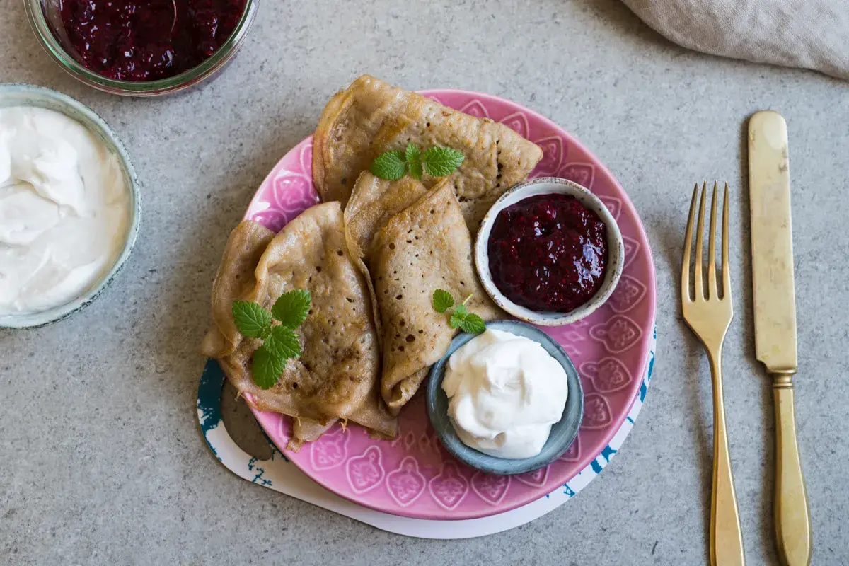 En tallrik med glutenfria pannkakor, serverade med sylt och grädde. En gyllene gaffel och kniv ligger bredvid.