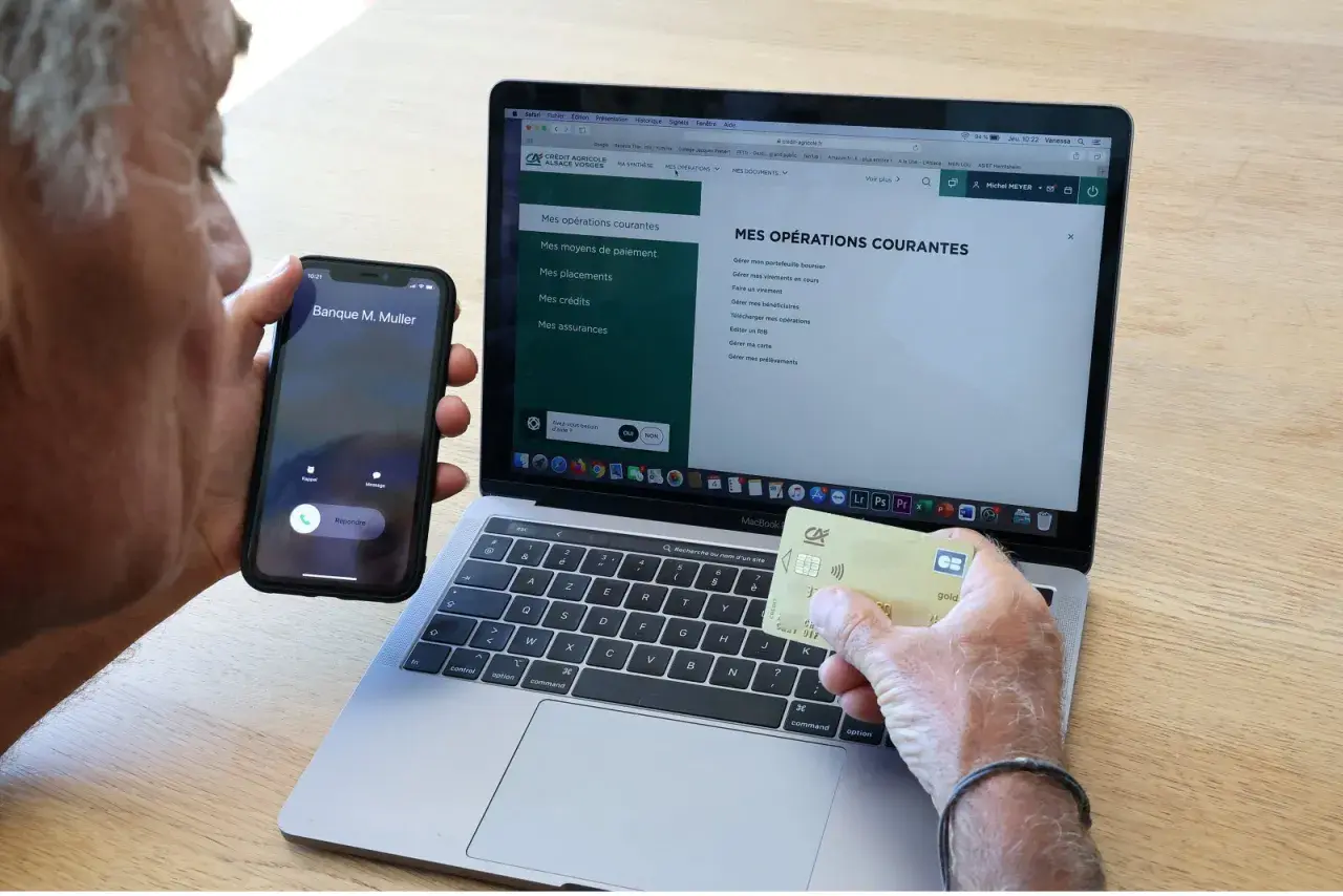 Un homme tient un téléphone et une carte bancaire, devant un ordinateur affichant le site du Crédit Agricole. Il cherche peut-on annuler un paiement par carte bancaire sur internet.