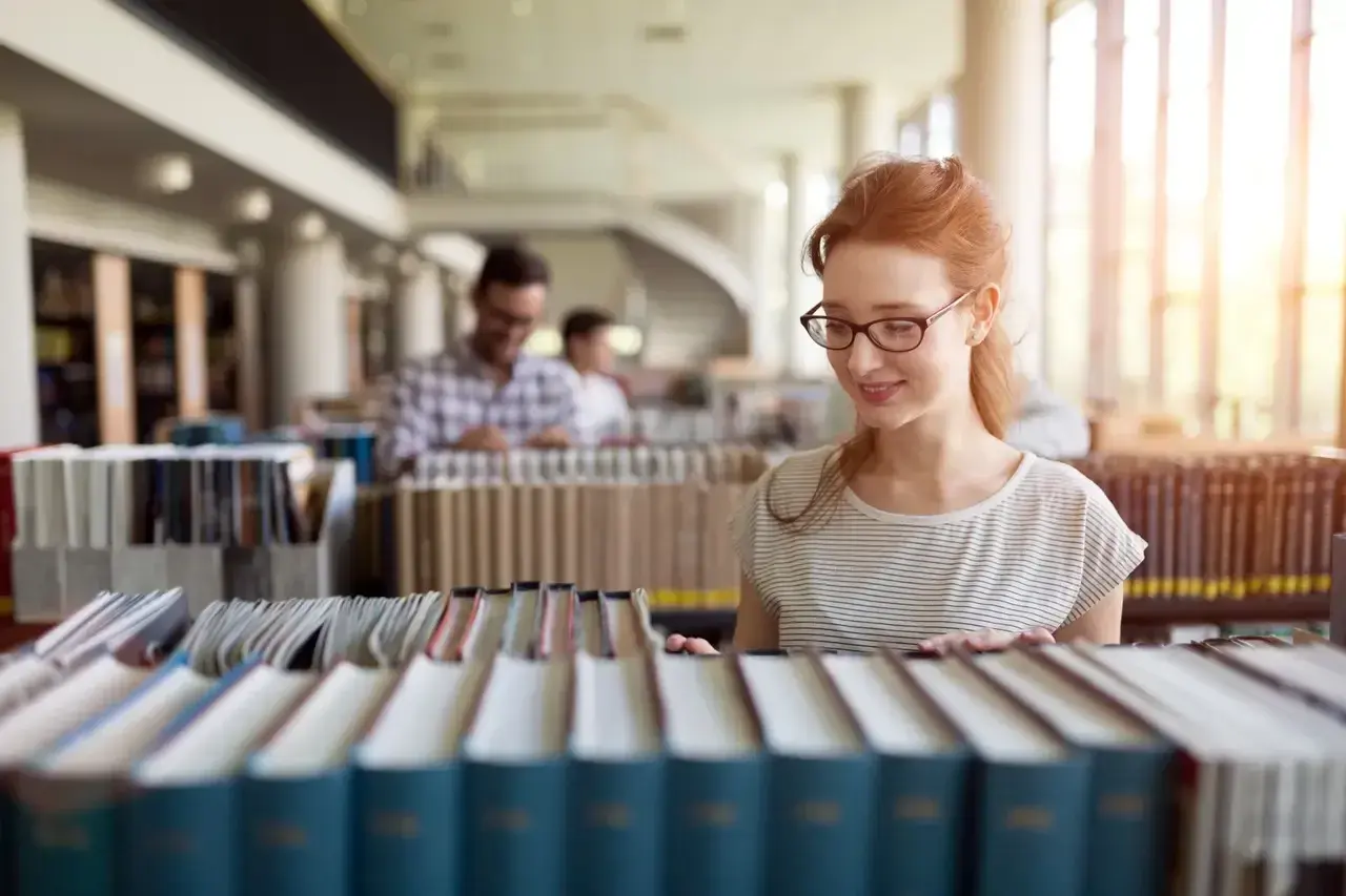 Studentka z rudymi włosami w okularach przegląda książki w bibliotece, szukając materiałów do prawo studia.