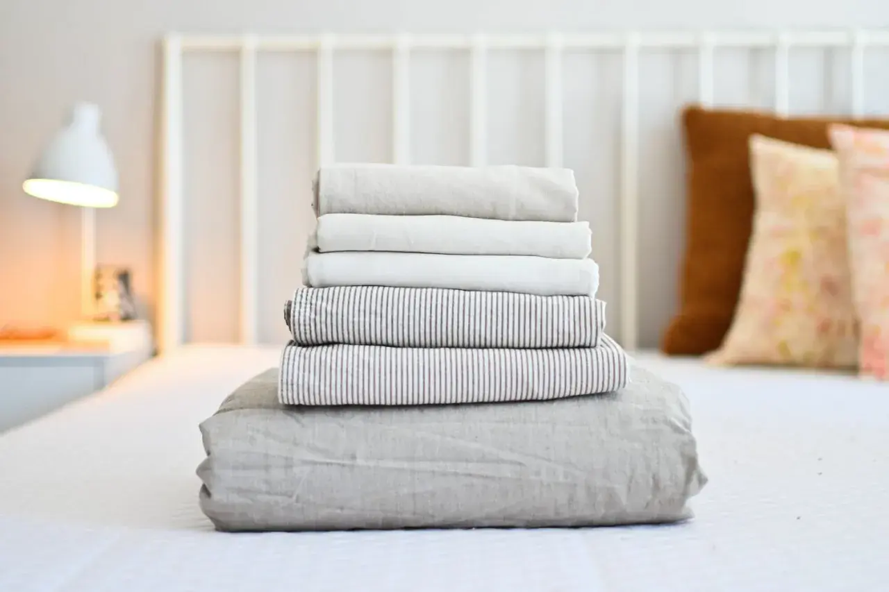 Neatly folded stack of clean bed sheets on a white bed, ready for how to wash bed sheets in washing machine.