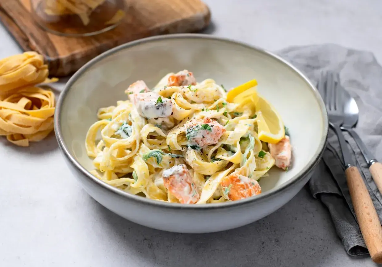 Pyszny makaron z łososiem w kremowym sosie, udekorowany cytryną i natką pietruszki. Obok leżą surowe tagliatelle.