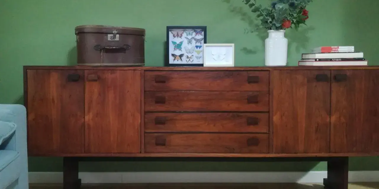 Mobile antico in legno scuro, con cassetti e ante. Sopra, una valigia vintage, quadri di farfalle e un vaso con fiori. Guida su come pulire un mobile antico.