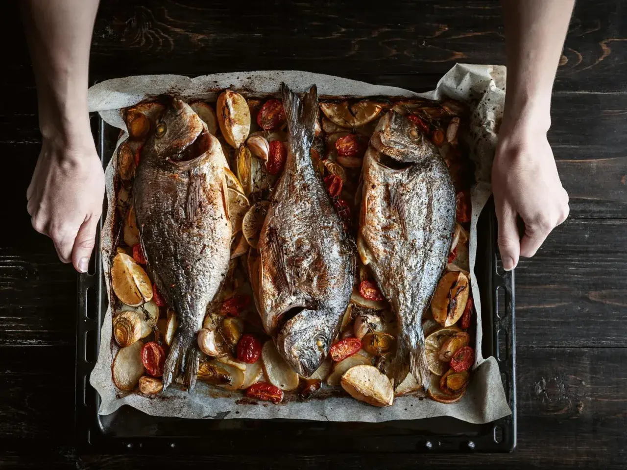 Tres pescados dorados al horno, acompañados de rodajas de limón, tomates cherry y ajos, listos para servir.