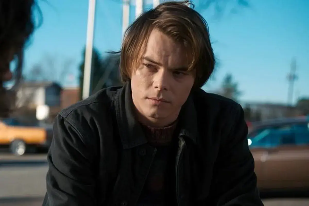 Charlie Heaton w ciemnej kurtce i swetrze, zamyślony, na tle błękitnego nieba i samochodów.
