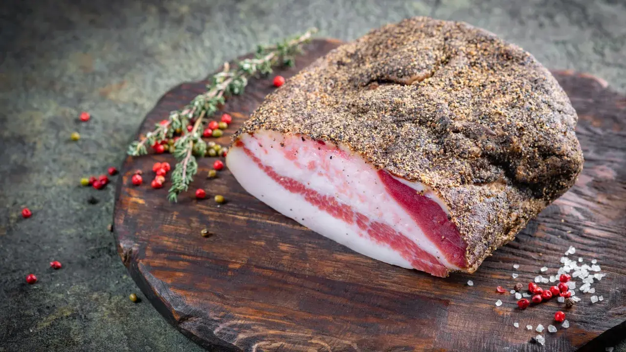 Pancetta co to? Wędzony kawałek boczku z widocznymi warstwami tłuszczu i mięsa, przyprawiony pieprzem, leży na drewnianej desce z ziołami i ziarnami pieprzu.