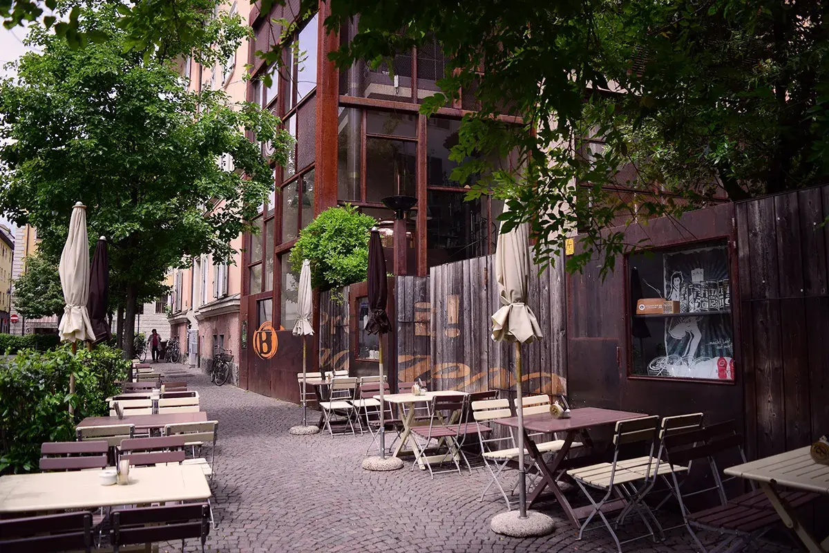 Tavoli all'aperto e ombrelloni di fronte al Batzen Bräu, con un'architettura industriale e un'atmosfera rilassata.